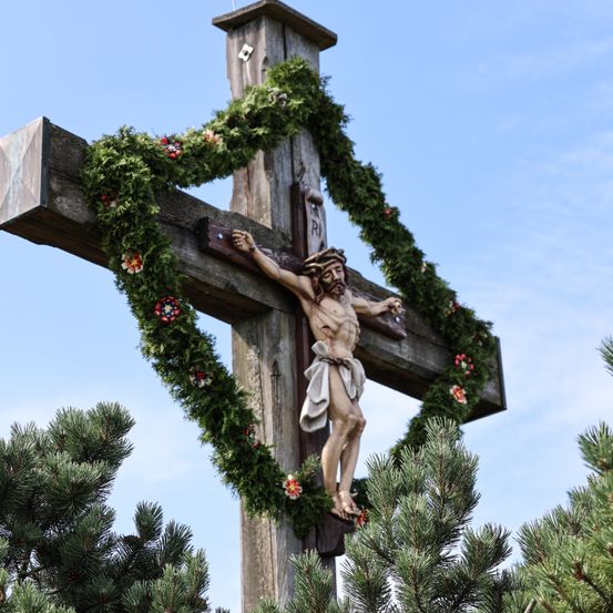 Ein hölzerne Kreuz mit dem gekreuzigten Jesus Christus ist von einem Kranz aus grünen Blättern umgeben. Das Kreuz steht vor einem blauen Himmel.