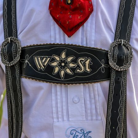 Ein Nahaufnahme eines traditionellen deutschen Outfits mit einem schwarzen Ledergürtel, auf dem 'West' in Gold gestickt ist. Der Gürtel ist mit aufwendigen Blumendesigns und silbernen Schnallen verziert. Ein roter Schal mit goldener Stickerei ist um den Hals gebunden.