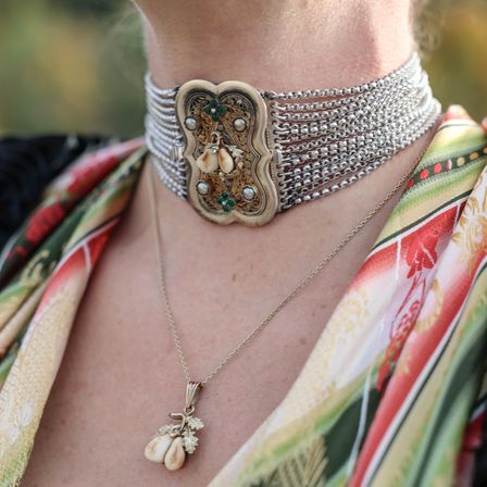 Eine Frau trägt eine silberne Halskette mit einer Schnalle, die grüne und perlenartige Designs aufweist, und einen silbernen Anhänger mit einem Blatt und zwei weißen Perlen.