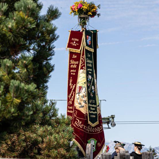 Ein Banner mit einer Blumenanordnung oben ist an einem Pfahl aufgehängt. Es hat rote und grüne Farben und etwas Text. Zwei Menschen mit Hüten stehen hinter dem Pfahl.