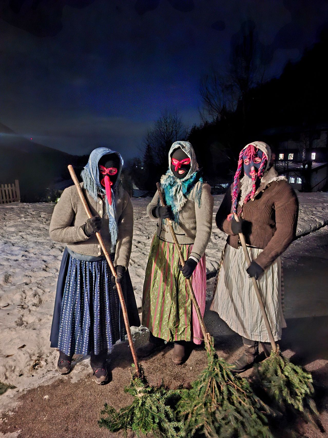Drei vermummte Personen in traditioneller Kleidung stehen bei Nacht im Schnee und halten Stöcke und Grünpflanzen. Sie scheinen ein Ritual durchzuführen.