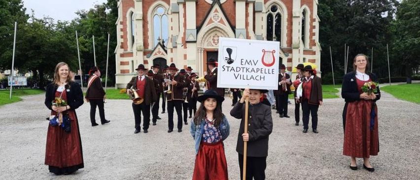 Gruppe von Menschen in traditioneller Kleidung, die mit einem Banner vor einer Kirche marschieren.