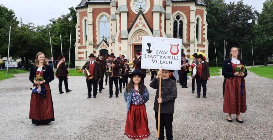 Gruppe von Menschen in traditioneller Kleidung, die mit einem Banner vor einer Kirche marschieren.
