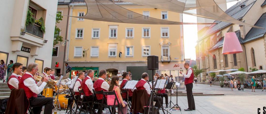 Ein Straßenorchester spielt unter einem großen Baldachin vor einem gelben Gebäude mit Glockenturm. Eine Frau steht neben den Musikern, und es gibt Leute, die in der Gegend herumlaufen.