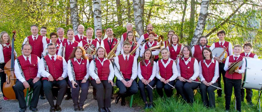 Eine große Gruppe von Musikern in roten Westen und weißen Hemden posiert für ein Foto in einem bewaldeten Gebiet. Sie halten verschiedene Instrumente.