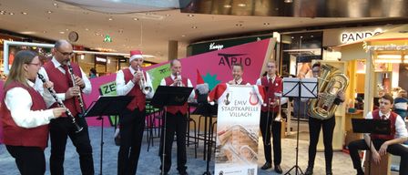 Eine Gruppe von Musikern, die rote Westen und Weihnachtsmützen tragen, spielt in einem Einkaufszentrum. Sie haben Trompeten, Posaunen und Tuben. Hinter ihnen steht ein Banner.