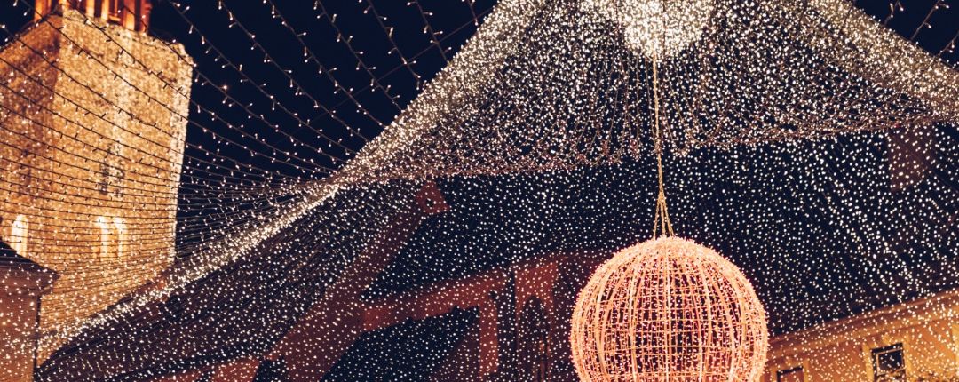 Eine festliche Szene mit einer großen beleuchteten Kugel, die von der Decke hängt, umgeben von funkelnden Lichterketten. Die Umgebung scheint ein nächtliches Outdoor-Event zu sein.