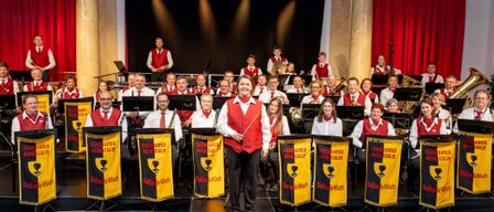 Eine Gruppe von Musikern in roten Westen und Krawatten steht auf der Bühne mit einem Banner. Eine Frau in der Mitte hält einen Taktstock. Hinter ihnen spielen andere Musiker Instrumente. Ein schwarzer Vorhang ist im Hintergrund.