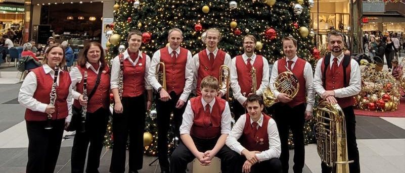 Eine Gruppe von Musikern posiert vor einem geschmückten Weihnachtsbaum in einem Einkaufszentrum. Der Baum ist mit roten, goldenen und silbernen Ornamenten geschmückt. Die Musiker tragen rote Westen und spielen Instrumente wie Trompeten und Klarinetten. Darüber hängt ein rotes Dekorationsstück.