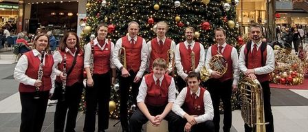 Eine Gruppe von Musikern posiert vor einem geschmückten Weihnachtsbaum in einem Einkaufszentrum. Der Baum ist mit roten, goldenen und silbernen Ornamenten geschmückt. Die Musiker tragen rote Westen und spielen Instrumente wie Trompeten und Klarinetten. Darüber hängt ein rotes Dekorationsstück.
