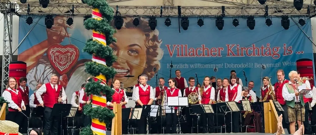 Eine Band tritt auf der Bühne auf, mit einem Banner hinter ihnen. Das Banner trägt den Text 'Villacher Kirchtag'. Die Bandmitglieder tragen rote Westen. Sie spielen verschiedene Instrumente. Die Bühne ist mit Weihnachtsbäumen geschmückt.