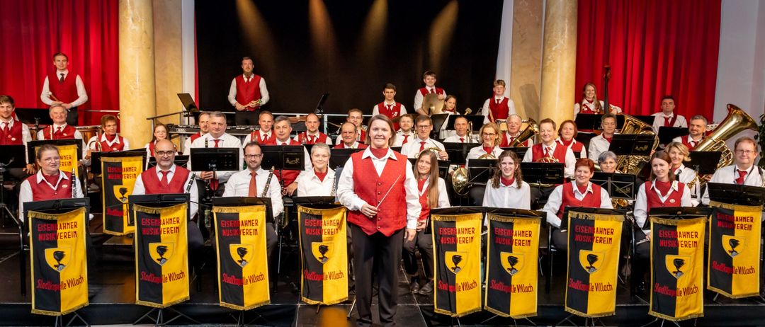 Eine Gruppe von Musikern tritt auf einer Bühne mit Musikinstrumenten auf. Die Person vorne hält einen Taktstock. Dahinter sitzen Leute auf Stühlen und spielen ihre Instrumente. Musikalische Banner stehen vor ihnen.