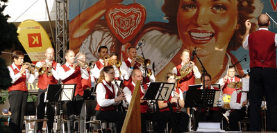Ein großes Orchester spielt auf der Bühne, mit Musikern, die Trompeten, Posaunen und andere Instrumente spielen. Ein großes Poster einer lächelnden Frau ist im Hintergrund zu sehen.