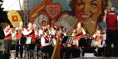 Ein großes Orchester spielt auf der Bühne, mit Musikern, die Trompeten, Posaunen und andere Instrumente spielen. Ein großes Poster einer lächelnden Frau ist im Hintergrund zu sehen.