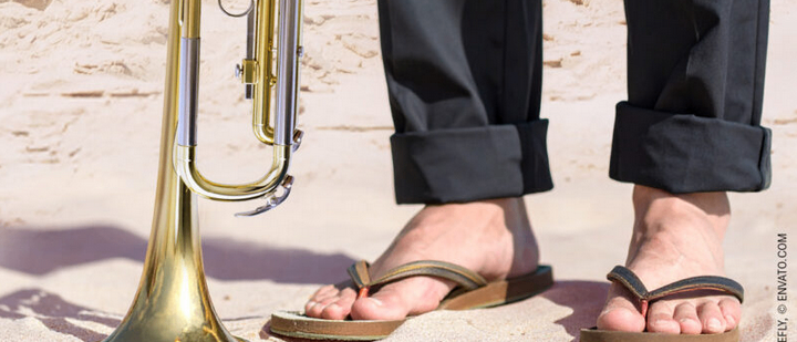 Eine Person in Sandalen steht barfuß auf einem sandigen Strand, mit einer Trompete, die vor ihnen auf dem Sand liegt. Die Person trägt schwarze Hosen und die Trompete ist golden.