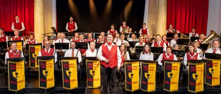 Eine Gruppe von Musikern, die rote Westen tragen und Musikinstrumente halten, steht auf einer Bühne. Sie lächeln und schauen in die Kamera. Hinter ihnen stehen andere Musiker, die verschiedene Instrumente spielen. Sie sind wahrscheinlich bei einem Konzert aufgetreten.