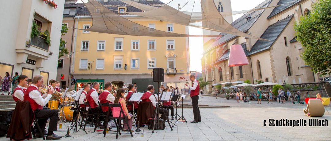 Ein Orchester spielt auf einem offenen Platz in der Stadt, mit einer Kirche im Hintergrund. Die Musiker sitzen unter einem Baldachin. Es gibt Menschen, die herumlaufen, und Gebäude sind im Hintergrund zu sehen.