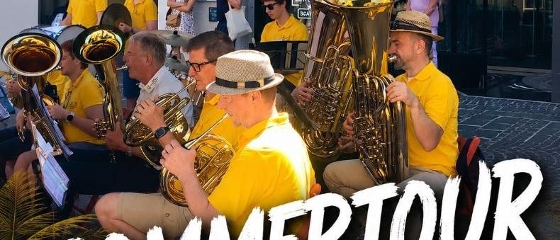 Ein Werbeplakat für die Villach Sommer Tour 2026. Es zeigt eine Blaskapelle bei einer Aufführung. Einige tragen Hüte, andere gelbe Hemden. Einige Leute schauen zu.