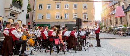 Ein Orchester spielt auf einem Stadtplatz vor einer Kirche mit Glockenturm, unter einem großen Baldachin.