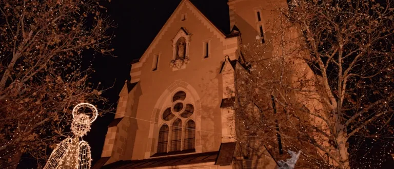 Ein Kirchgebäude mit Turm und Uhr ist bei Nacht beleuchtet. Über dem Eingang befindet sich eine Statue eines Heiligen.