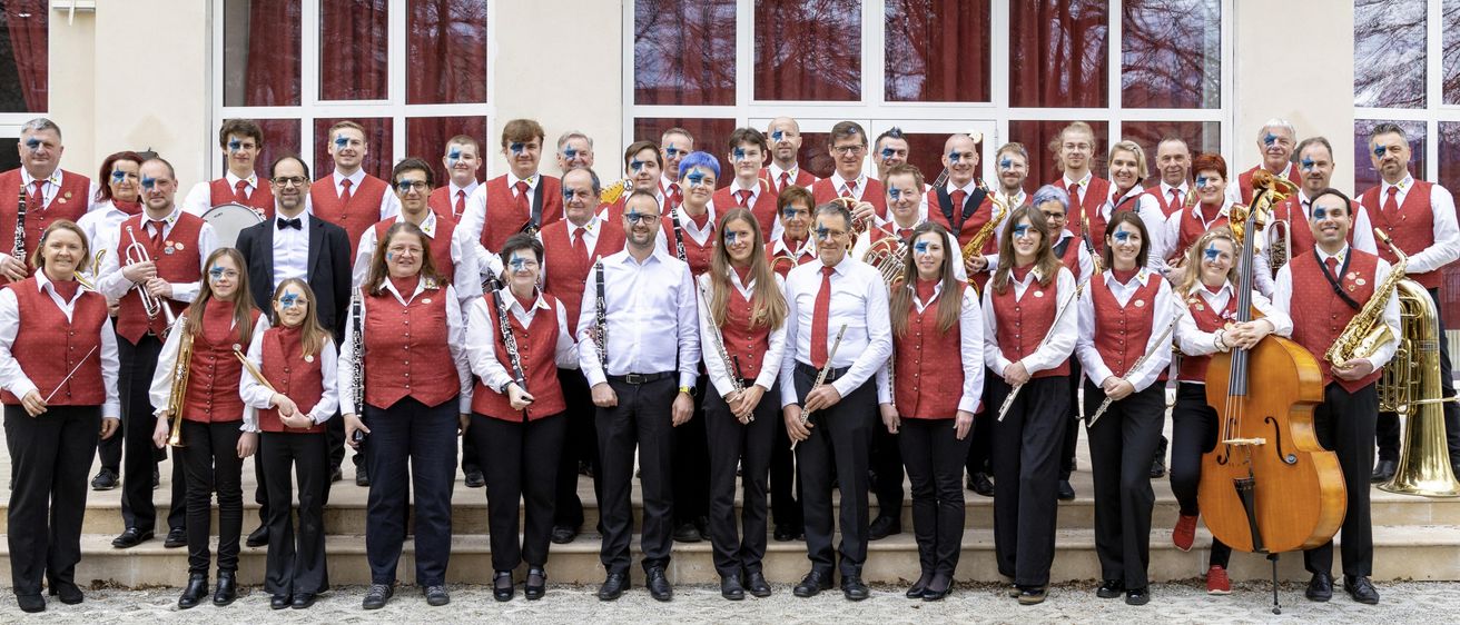 A group of musicians in red vests and face paint pose for a photo outside a building. They are holding instruments like flutes, saxophones, and trumpets.