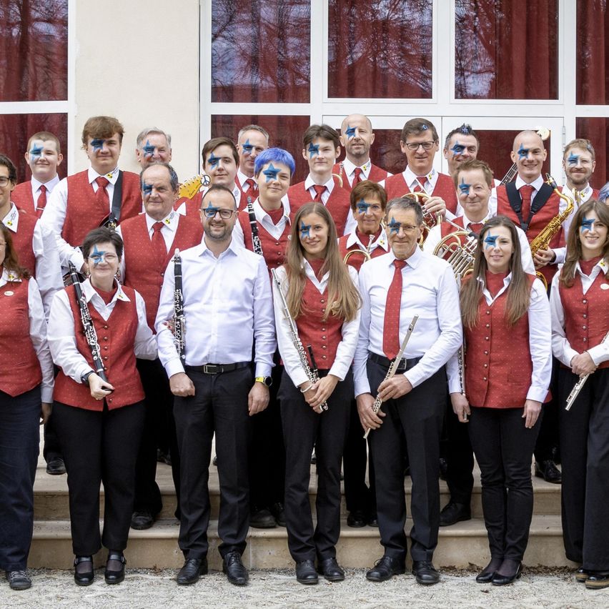 A group of musicians in red vests and face paint pose for a photo outside a building. They are holding instruments like flutes, saxophones, and trumpets.