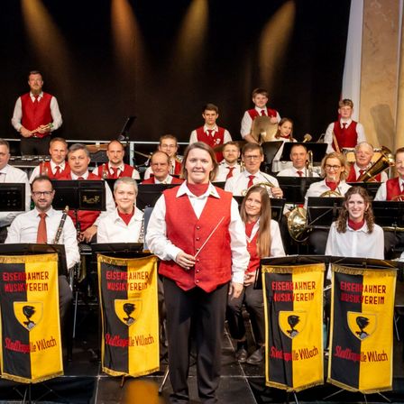 Eine Gruppe von Musikern in roten Westen und mit Instrumenten auf der Bühne, mit einer Frau, die vorne einen Taktstock hält.