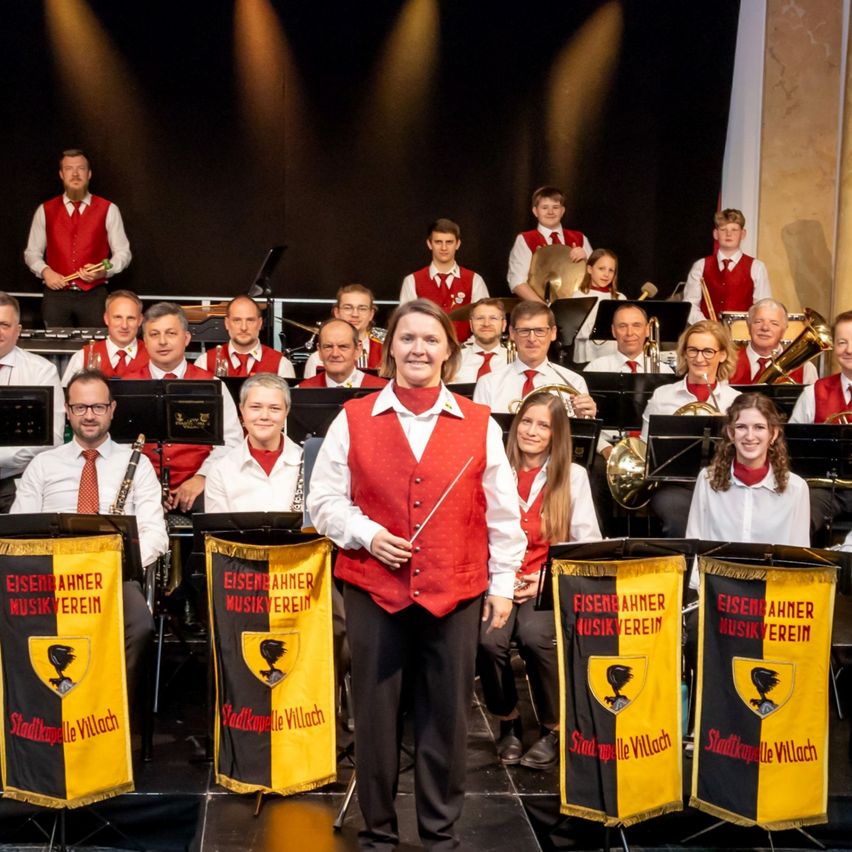 Eine Gruppe von Musikern in roten Westen und mit Instrumenten auf der Bühne, mit einer Frau, die vorne einen Taktstock hält.