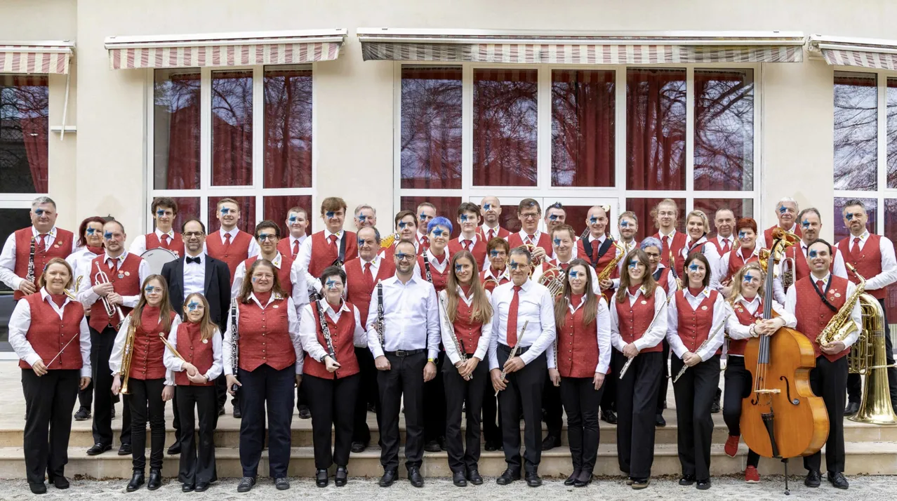 Eine Gruppe von Musikern in roten Westen und weißen Hemden posiert für ein Foto vor einem Gebäude mit großen Fenstern. Sie halten verschiedene Instrumente in den Händen und einige haben Gesichtsbemalung.