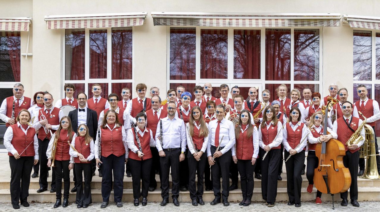 Eine Gruppe von Musikern in roten Westen und weißen Hemden posiert für ein Foto vor einem Gebäude mit großen Fenstern. Sie halten verschiedene Instrumente in den Händen und einige haben Gesichtsbemalung.
