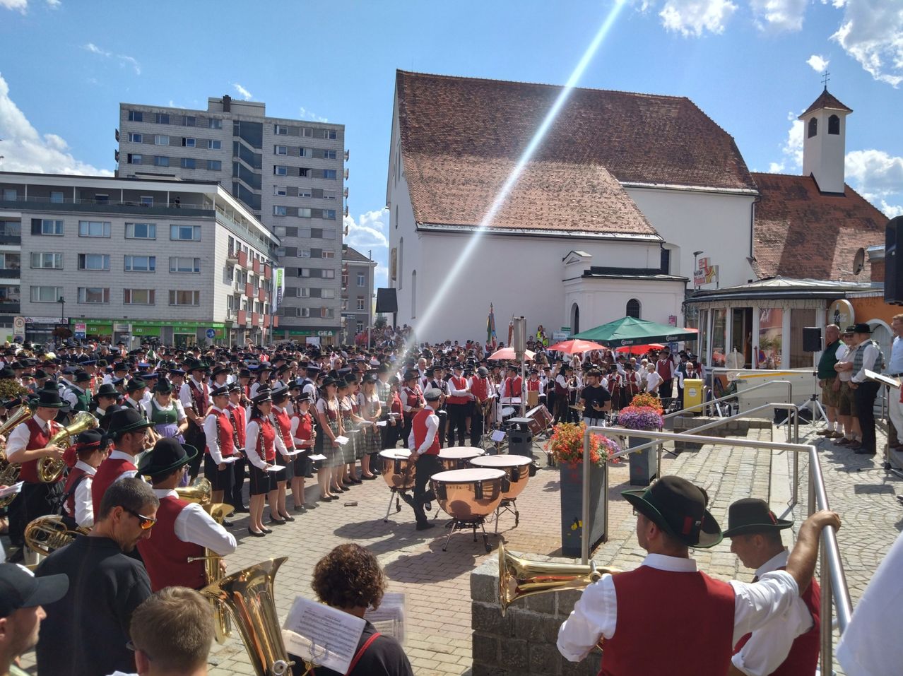 Eine große Gruppe von Musikern in traditioneller Kleidung spielt auf einem Freiluftgelände, mit einem Gebäude und Gebäuden im Hintergrund.