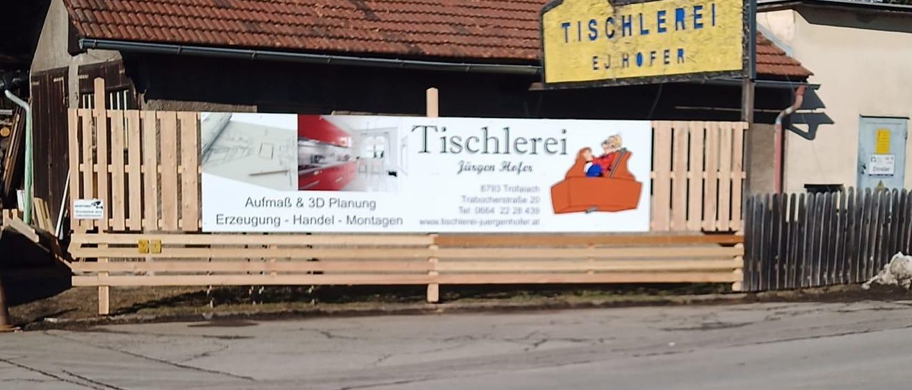 Ein Werbeschild für Tischlerei Jürgen Hofer steht vor einem Holzzaun. Dahinter befindet sich ein Gebäude mit einem Ziegeldach. Das Schild zeigt Möbelillustrationen.