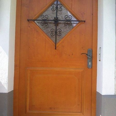 Eine hölzerne Tür mit einem metallischen kreuzförmigen Design und einem silbernen Griff ist an einer weißen Wand montiert.