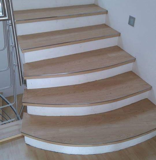 Eine Holztreppe mit mehreren Stufen, weißen Verkleidungen und einem silbernen Handlauf. Der Boden ist aus Holz und die Wand ist weiß.