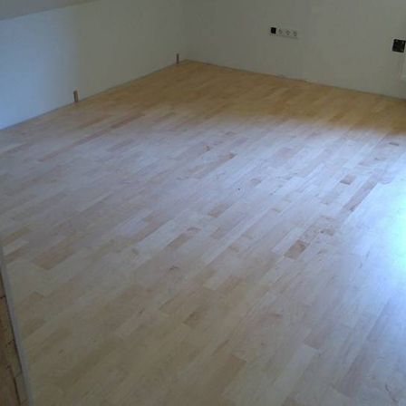 Ein leerer Raum mit weißen Wänden und Holzböden, beleuchtet von Sonnenlicht. Ein weißer Heizkörper ist an der Wand montiert.