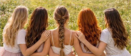 Five women sit in a flowery field, their backs turned. They are surrounded by flowers and greenery, with a house and mountain range in the distance. The words 'Zum Durchatmen und Wieder Aufblühen' are written above them.