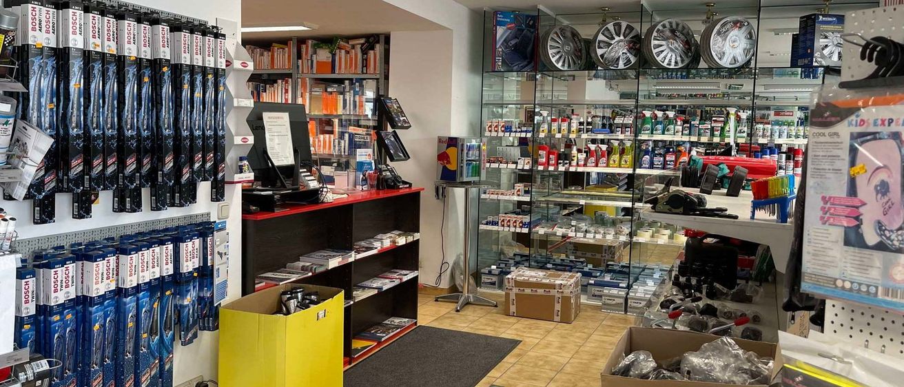 Ein gut organisierter Laden mit Regalen voller Bücher und Autoteilen, einem leuchtend gelben Beutel und einem schwarzen Teppich auf dem Boden. Ein Schreibtisch mit Computerbildschirm und anderen Gegenständen steht in der Mitte, und ein Glasvitrine mit Produkten ist rechts.