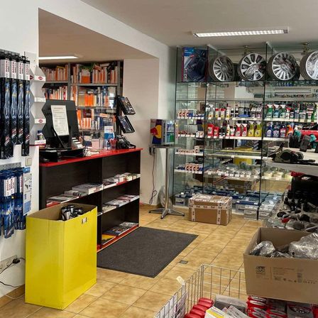 Ein Autoteileladen mit schwarzem Tresen und Regalen, einer Glasvitrine und einem gelben Mülleimer. Der Laden hat einen gefliesten Boden, Regale mit verschiedenen Artikeln und einen Monitor auf dem Tresen.