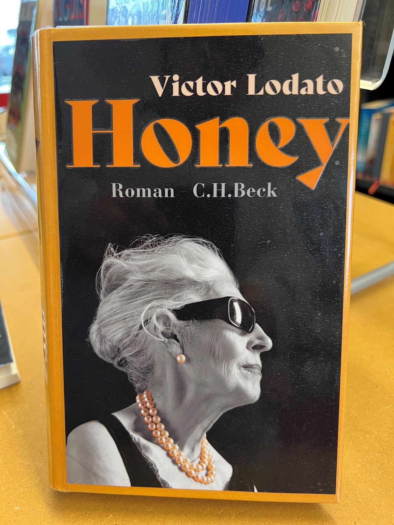 Ein Schwarz-Weiß-Foto einer älteren Frau mit grauem Haar, die eine Sonnenbrille, Perlen und ein ärmelloses Top trägt, vor schwarzem Hintergrund.