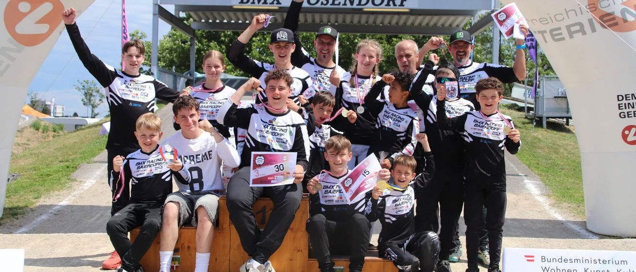 Eine Gruppe von BMX-Rennfahrern, sowohl jung als auch erwachsen, feiert unter einem weißen Bogen mit einem Banner, das BMX-Osendorf liest. Sie halten Medaillen und posieren für ein Foto.