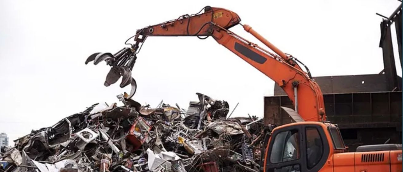 Ein oranger Bagger hebt Schrott in einem Schrottplatz, mit einem Gebäude im Hintergrund.