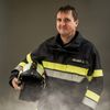 Ein Feuerwehrmann steht mit einem Helm, lächelt und trägt eine schwarze und gelbe Uniform mit 'KELLNER J' auf der Brust.