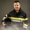 Ein lächelnder Mann in einem Feuerwehrmann-Uniform hält einen Helm, mit den Worten Rodler Ch auf seiner Jacke. Er steht vor einem grauen Hintergrund.