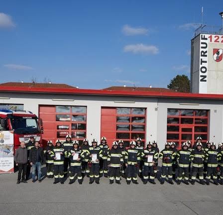 Eine Gruppe von Feuerwehrleuten steht vor einem Feuerwehrhaus, mit einem Feuerwehrauto davor, an einem sonnigen Tag.