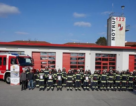 Eine Gruppe von Feuerwehrleuten steht vor einem Feuerwehrhaus, mit einem Feuerwehrauto davor, an einem sonnigen Tag.