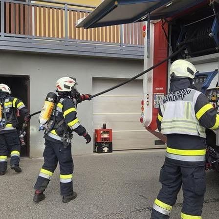 Feuerwehrleute in Uniform mit Helmen bereiten sich darauf vor, ein Gebäude mit einer Feuerwehrspritze zu betreten. Das Fahrzeug steht vor ihnen.