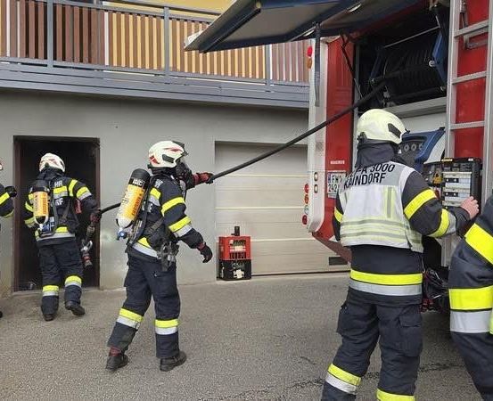 Feuerwehrleute in Uniform mit Helmen bereiten sich darauf vor, ein Gebäude mit einer Feuerwehrspritze zu betreten. Das Fahrzeug steht vor ihnen.
