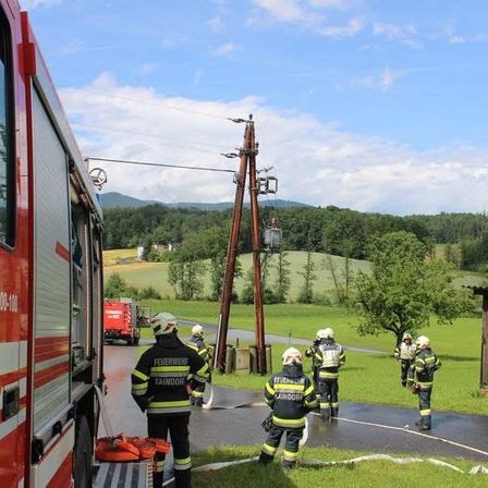 Feuerwehrleute stehen in Schutzkleidung auf einer Straße mit einem Feuerwehrauto im Hintergrund.