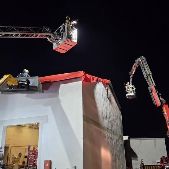 Feuerwehrleute in Helmen und Gurten benutzen einen Kran, um eine rote Abdeckung vom Dach eines Gebäudes bei Nacht zu entfernen.