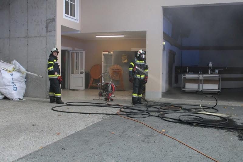 Zwei Feuerwehrleute stehen draußen mit Schutzausrüstung, einer hält einen Schlauch, und ein Feuerlöscher im Hintergrund.